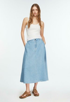 Kobieta stojąca z rękami w kieszeniach, ubrana w białą koszulkę na ramiączkach, jasnoniebieską dżinsową spódnicę midi i brązowe sandały z odkrytymi palcami na białym tle.