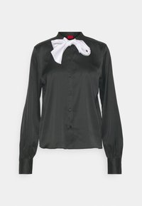 Blusa nera di satin con maniche lunghe, chiusura frontale con bottoni e un accento di fiocco bianco al colletto, caratterizzata da una texture liscia e lucida.
