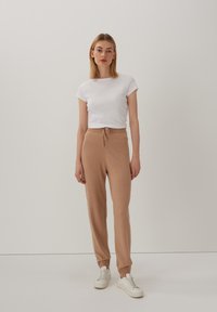 Leichtes, kurzärmliges weißes Oberteil und tanfarbene Jogginghose mit Schnürzug. Die Jogginghose hat eine weiche Textur und einen lockeren Sitz. Getragen mit weißen Sneakers.