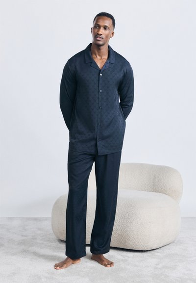 Egy férfi áll mezítláb, sötétkék, gombos, hosszú ujjú felsőt és nadrágot viselve egy fehér, texturált karosszék előtt, szürke szőnyegen.