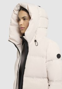 Puffy white jacket with a large hood, black accents, and zip closure. Features a textured fabric and a logo patch on the sleeve.
