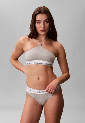 Femme portant un bralette gris Calvin Klein à col halter et un slip assorti, debout avec une main sur la hanche sur un fond gris uni.