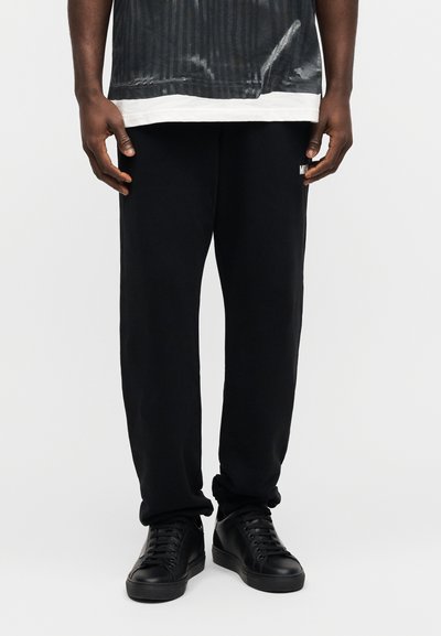 Άνδρας που φοράει μαύρο casual παντελόνι και μαύρα sneakers με κορδόνια, στέκεται μπροστά σε απλό λευκό φόντο.