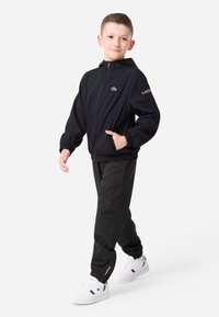 Μαύρο τζάκετ με φερμουάρ και κουκούλα, ελαστικούς καρπούς και τσέπες, σε συνδυασμό με μαύρες φόρμες και άσπρα αθλητικά παπούτσια με χρωματικές λεπτομέρειες.