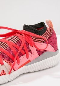 Sportovní obuv s pleteným svrškem v růžové a černé barvě s červenými prvky, s polstrovaným límcem a texturovanou bílou podrážkou pro lepší přilnavost.