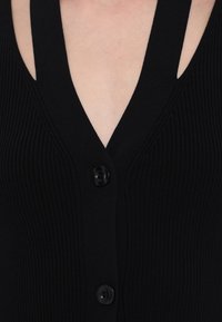 Gros plan sur un cardigan noir côtelé avec deux boutons visibles et un profond décolleté en V laissant apparaître la peau en dessous.