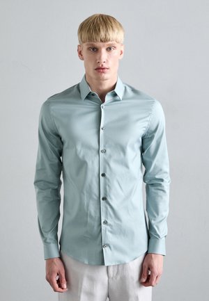 Light blue long-sleeve shirt made of smooth fabric, featuring a pointed collar, front buttons, and fitted design, worn with light-colored trousers.