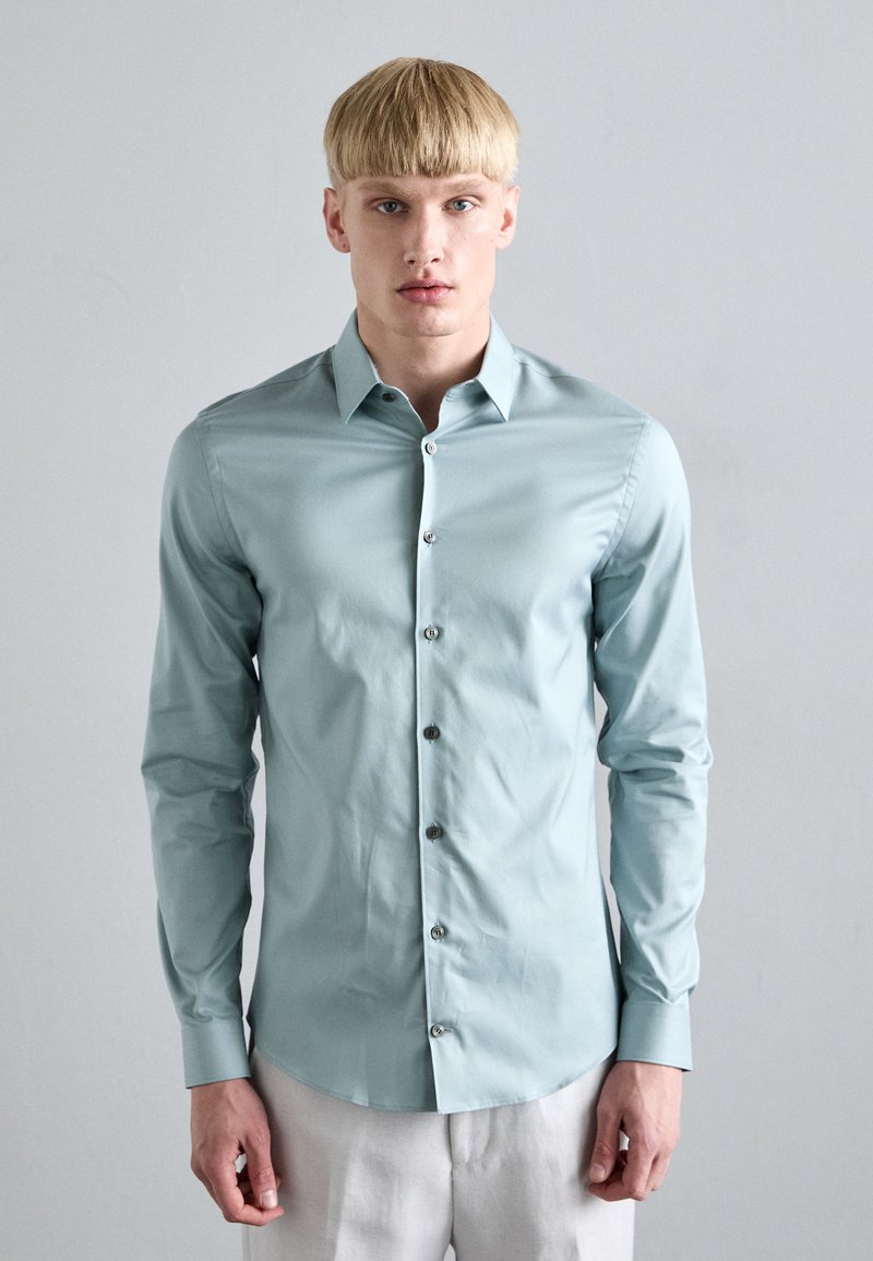 Camisa de manga larga azul claro hecha de una tela suave, con cuello puntiagudo, botones en la parte delantera y un diseño ajustado, combinada con pantalones de color claro.