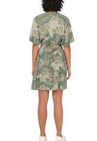 Vestido floral verde con mangas cortas, cintura anudada y patrones de hojas en beige y blanco, hecho de un tejido ligero. Corte casual.