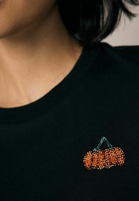 T-shirt noir avec un petit design brodé de trois citrouilles orange, rehaussé de tiges vertes, situé près de l'encolure.