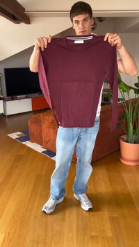 Jauns vīrietis džinsos un sporta apavos tur tumši sarkanu garām piedurknēm krekliņu gaišā viesistabā ar koka grīdu un augiem.
