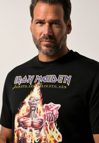 Černé bavlněné tričko s barevným grafickým potiskem kostlivce v plamenech a textem "Iron Maiden" nahoře v tučném žlutém písmu.