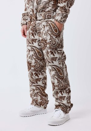 Persoon draagt bruine en beige broek met paisley patroon en bijpassend jasje met witte sneakers, handen in de zakken, tegen een effen achtergrond.