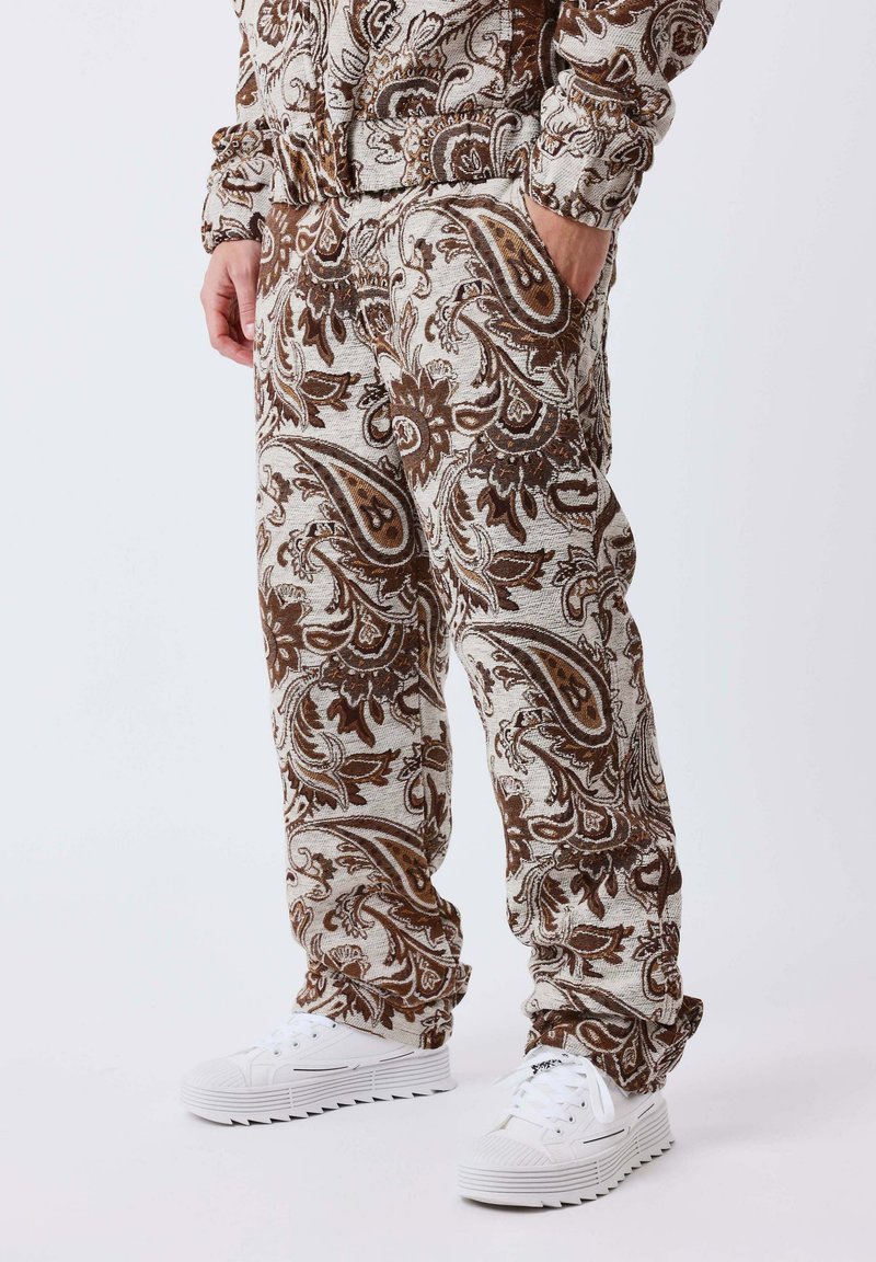 Personne portant un pantalon marron et beige à motif paisley et une veste assortie avec des baskets blanches, mains dans les poches, sur un fond uni.
