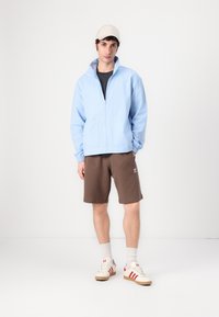Light blue zip-up jacket with elastic cuffs, black shirt, brown shorts, and cream sneakers with red accents; worn with a beige cap and white socks.