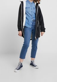 Czarna kurtka z kapturem i białym wykończeniem, niebieska koszula dżinsowa, niebieskie jeansy typu cropped oraz granatowe sneakersy za kostkę z białymi podeszwami.