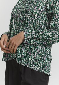 Blusa floreale con motivi verdi, viola e bianchi, caratterizzata da dettagli arricciati, maniche lunghe e una vestibilità ampia. Indossata con pantaloni scuri.