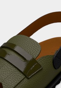 Πράσινο δερμάτινο σανδάλι με υφή, που διαθέτει λουρί με γυαλιστερή λεπτομέρεια και καφέ πάτο. Περιλαμβάνει διακοσμητικό μοτίβο με κοψίματα.