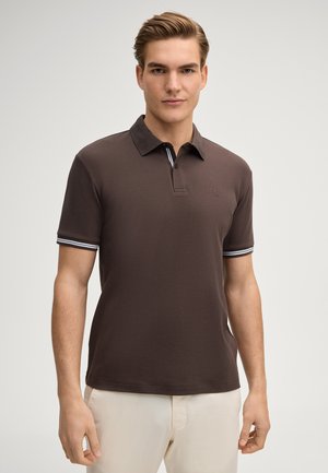 Mężczyzna w ciemnobrązowej koszulce polo z krótkim rękawem, z paskami na krawędziach rękawów, oraz w beżowych spodniach, stojący na jednolitym tle.