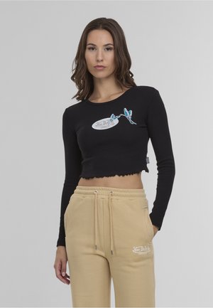 Žena oblečená v čiernom crop top s dlhým rukávom s modrým motýľovým motívom a béžových teplákoch so sťahovacou šnúrkou a logom Von Dutch.