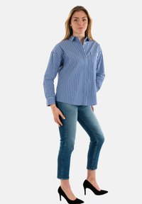 Camisa de manga larga a rayas azules y blancas con cuello de botones, combinada con jeans ajustados y zapatos de tacón alto negros.