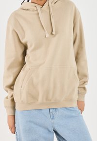 Sudadera beige con capucha y cordones, bolsillo frontal y puños acanalados, combinada con jeans de mezclilla azul claro. Tejido suave y texturizado.