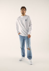 Cămașă albă cu mâneci lungi și un mic logo albastru, asortată cu blugi evazați deschis, rupți, și adidași albi, stând în fața unui fundal neutru.