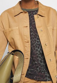 Tan denim jacket with button closures, paired with a patterned top featuring shades of green and black. Black and olive handbag displayed.