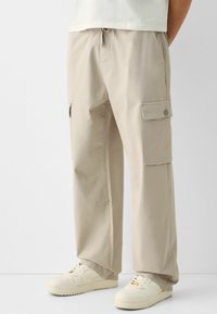 Beżowe spodnie cargo o luźnym kroju, z bocznymi kieszeniami i regulowanym paskiem. Noszone z białymi trampkami, które mają teksturowany materiał i czysty design.