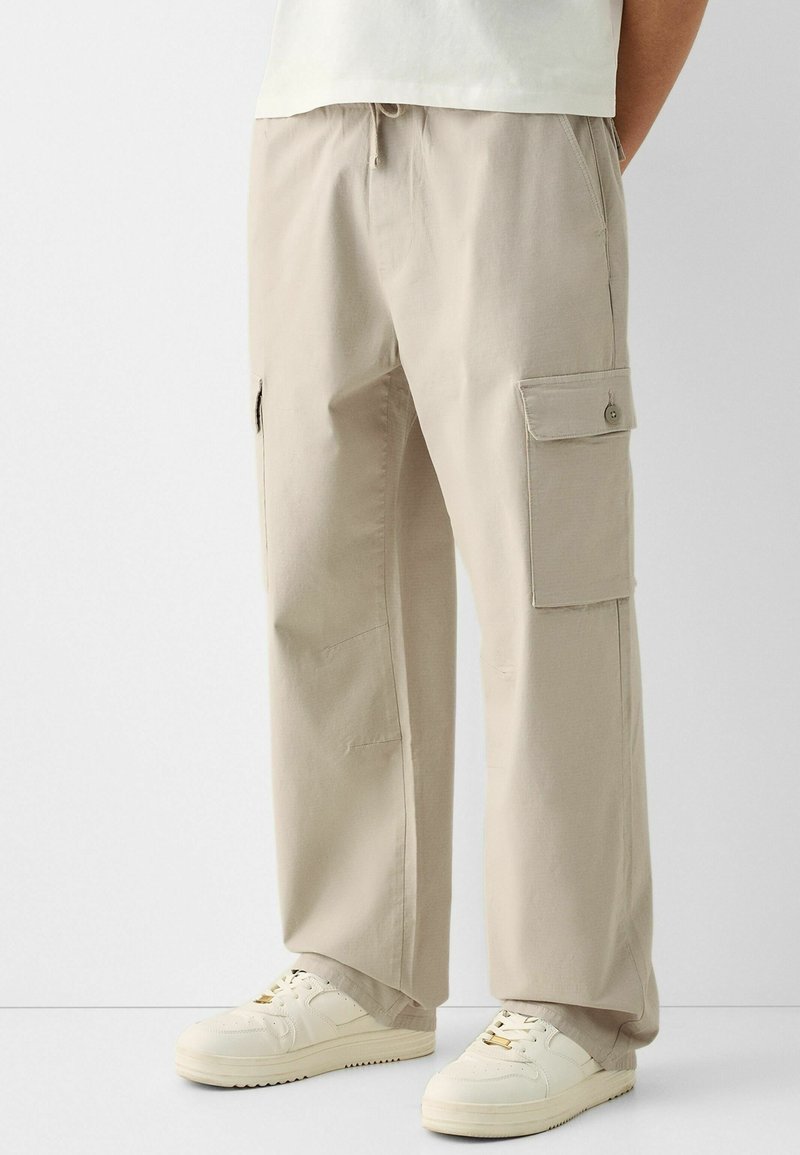Beżowe spodnie cargo o luźnym kroju, z bocznymi kieszeniami i regulowanym paskiem. Noszone z białymi trampkami, które mają teksturowany materiał i czysty design.