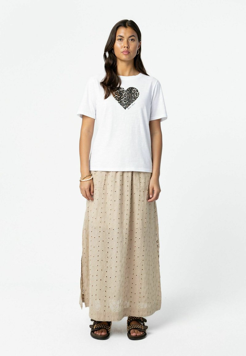 Femme debout de face portant un t-shirt blanc avec un cœur léopard imprimé, une jupe longue beige à œillets, des sandales noires cloutées et des bracelets.
