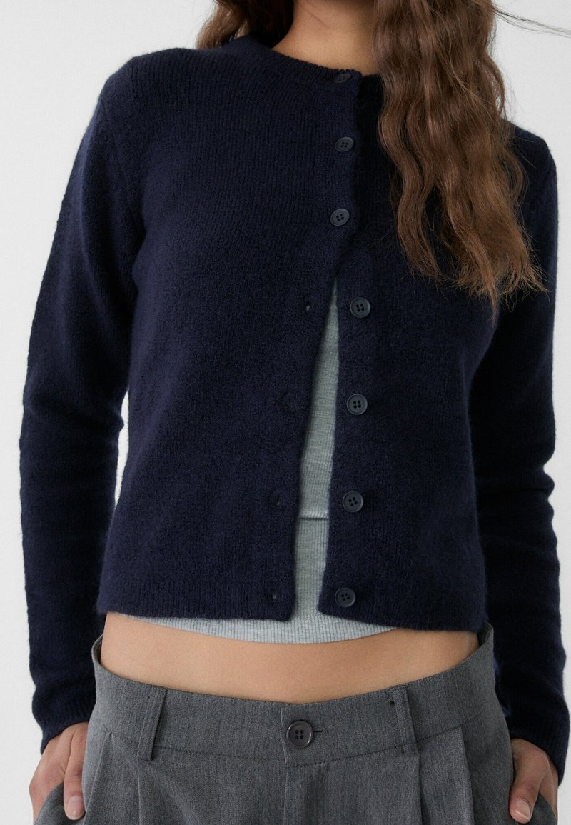 Femme portant un cardigan bleu marine boutonné par-dessus un top court gris et un pantalon gris taille haute, montrant le ventre et une partie des mains.