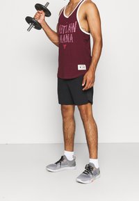 Bordeauxowa bluzka na ramiączkach z napisem "OUTLAW MANA", czarne szorty, szare buty sportowe z akcentami oraz hantle w jednej ręce. Białe tło.