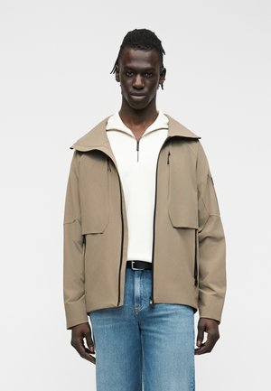 Mand med mørk hud og flettet hår iført en beige jakke over en hvid lynlåstrøje og blå jeans, stående foran en ensfarvet baggrund.