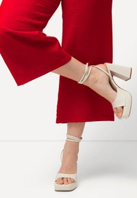 Witte hoge hakken sandalen met een chunky plateau, enkelbandjes en open teen. De voet is gepositioneerd tegen een rode stof achtergrond.