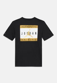 T-shirt nera con un logo rettangolare dorato e bianco di Jordan sulla schiena che recita "Authentic Jordan by Nike" e presenta una silhouette Jumpman dorata.