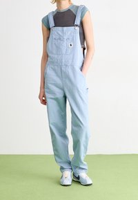 Hellblaue Jeans-Trägerhosen mit verstellbaren Trägern, einer Vorder tasche und geraden Beinen. Getragen über einem grauen und türkisfarbenen, kurzärmligen Hemd.