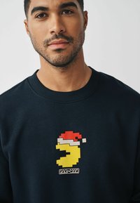 Felpa navy con una grafica di Pac-Man gialla che indossa un cappello di Babbo Natale rosso. Il design include elementi in pixel art e un colletto a coste.