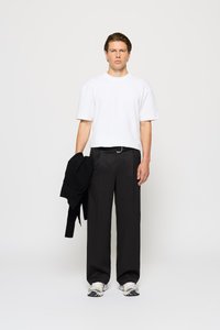 White cotton T-shirt, black tailored trousers, and gray sneakers. A black sweater is draped over his left arm. Simple, modern design.