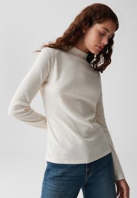 Langärmliges, cremefarbenes Oberteil aus geripptem Stoff mit gesprenkelten Mustern; hat einen hohen Halsausschnitt und eine figurbetonte Silhouette. Kombiniert mit dunklen Jeans.