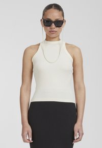 Top sin mangas de color beige claro con un cuello alto, combinado con una falda negra texturizada. Los accesorios incluyen gafas de sol negras y una cadena de perlas.