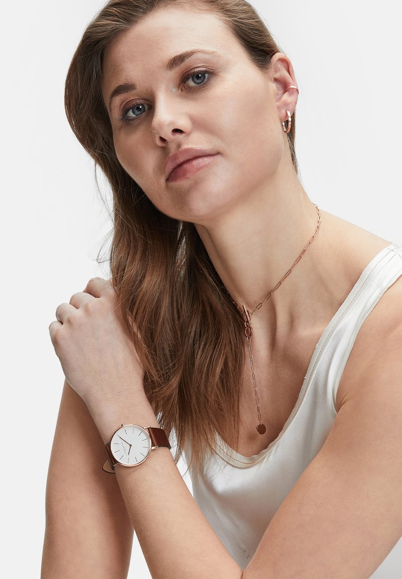 Braunes Lederarmbanduhr mit einem weißen Zifferblatt, minimalistisches Design, dünne Zeiger und roségoldene Akzente. Das Model trägt ein weißes Oberteil und mehrschichtige Halsketten.