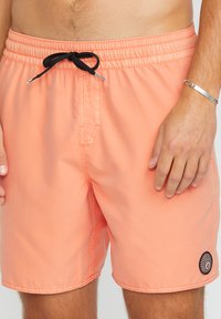 Pfirsichfarbene Badeshorts aus leichtem Material, mit einem elastischen Bund und schwarzem Kordelzug sowie einem runden Logoschild am Saum.