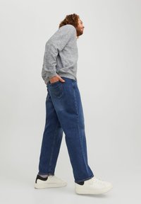 Grijze gebreide sweatshirt, losse blauwe jeans met achterzak en witte sneakers met zwarte accenten. Model in zijprofiel tegen een simpele achtergrond.