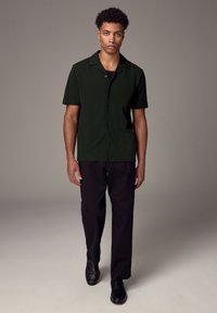 Camicia a maniche corte di colore verde scuro con colletto a punta, abbottonata sul davanti, abbinata a pantaloni neri e scarpe nere, sfondo semplice.
