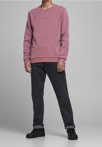 Felpa rosa con scollo rotondo e polsini a costine, abbinata a jeans grigi scuri a sigaretta e sneakers nere. La felpa presenta un delicato dettaglio testuale.