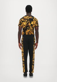 Black outfit featuring a short-sleeve shirt with a gold baroque pattern and black pants with gold side stripes, paired with black shoes.