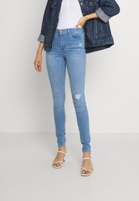 Hellblaue Skinny Jeans mit hoher Taille, die im Kniebereich abgerissen sind. Kombiniert mit einem weißen Oberteil und einer Jeansjacke, getragen mit wei�ßen Sandalen.