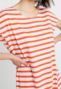 Pasiasta koszulka z szerokimi rękawami, z poziomymi pomarańczowymi i białymi paskami na lekkim materiale. Okrągły dekolt i luźny fason.