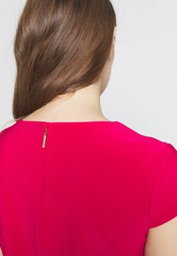 Fuchsie blůza s krátkými rukávy. Hladký výstřih na zádech, s diskrétním zipem a malým kovovým táhlem. Hladká textura.